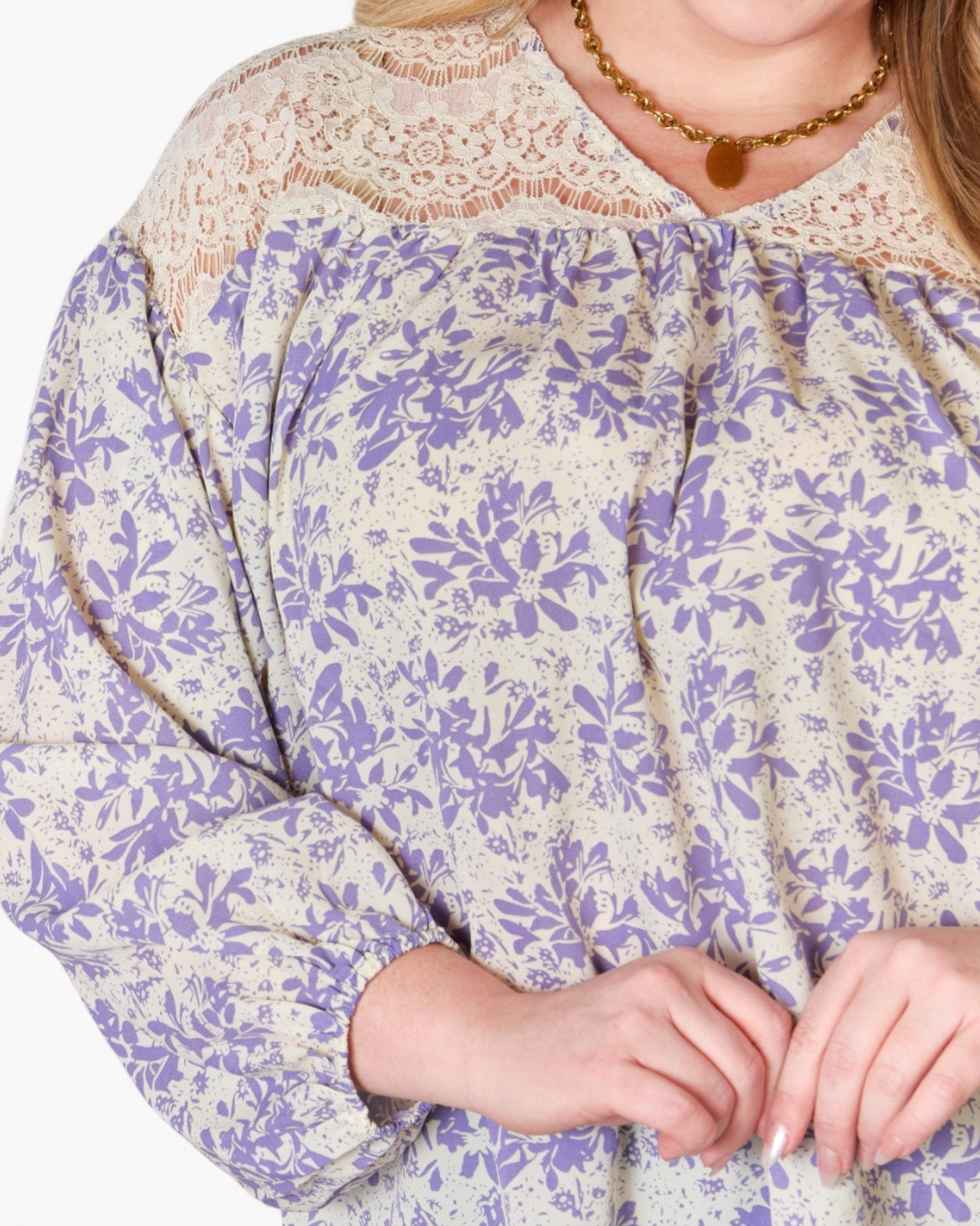 A close-up of a woman wearing a loose blouse with a lavender floral print on a cream background, featuring a beige lace yoke and a gold necklace.