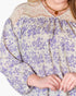 A close-up of a woman wearing a loose blouse with a lavender floral print on a cream background, featuring a beige lace yoke and a gold necklace.