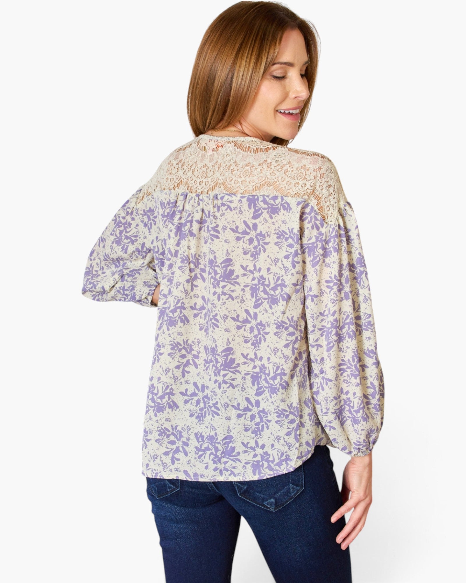 A woman wearing a cream-colored blouse with a purple floral pattern, featuring a beige lace yoke and puff sleeves, paired with dark blue jeans, shown from a three-quarter back view against a white studio background.