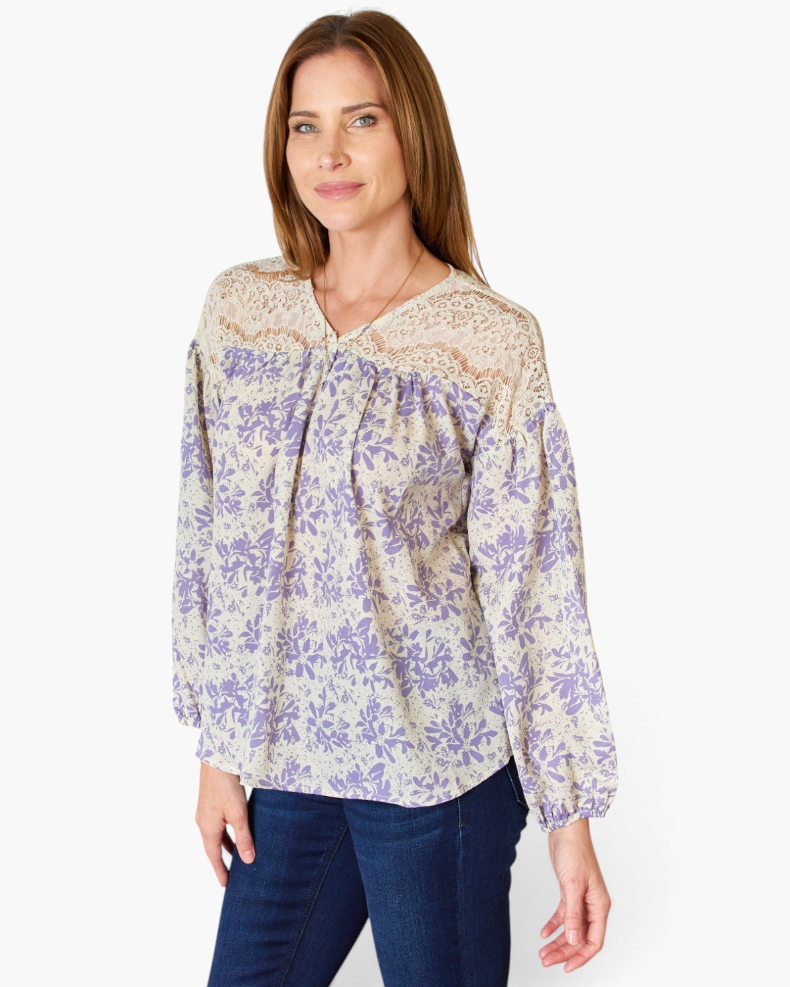 A woman wearing a long-sleeve blouse with a purple floral print on a cream background and a beige lace V-neck collar, paired with dark blue jeans, against a plain white background.