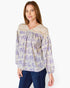 A woman wearing a long-sleeve blouse with a purple floral print on a cream background and a beige lace V-neck collar, paired with dark blue jeans, against a plain white background.