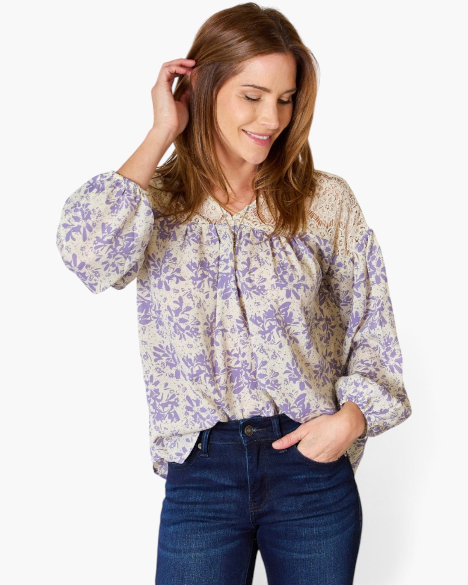 A woman wearing a beige long-sleeve blouse with a purple floral pattern and dark blue jeans, posing against a light gray background.