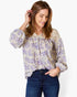 A woman wearing a beige long-sleeve blouse with a purple floral pattern and dark blue jeans, posing against a light gray background.