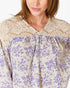A close-up of a woman wearing a blouse with a cream lace yoke and a purple floral print on a cream base.