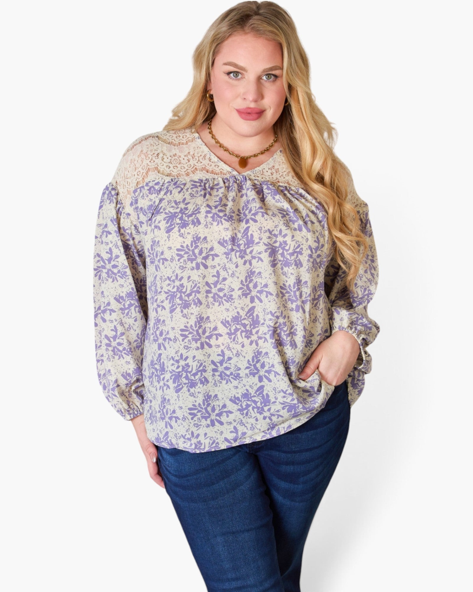 A woman modeling a long-sleeve blouse with a purple floral print on a cream background, featuring a lace-trimmed round neckline and gathered front, paired with dark blue jeans against a light gray studio background.