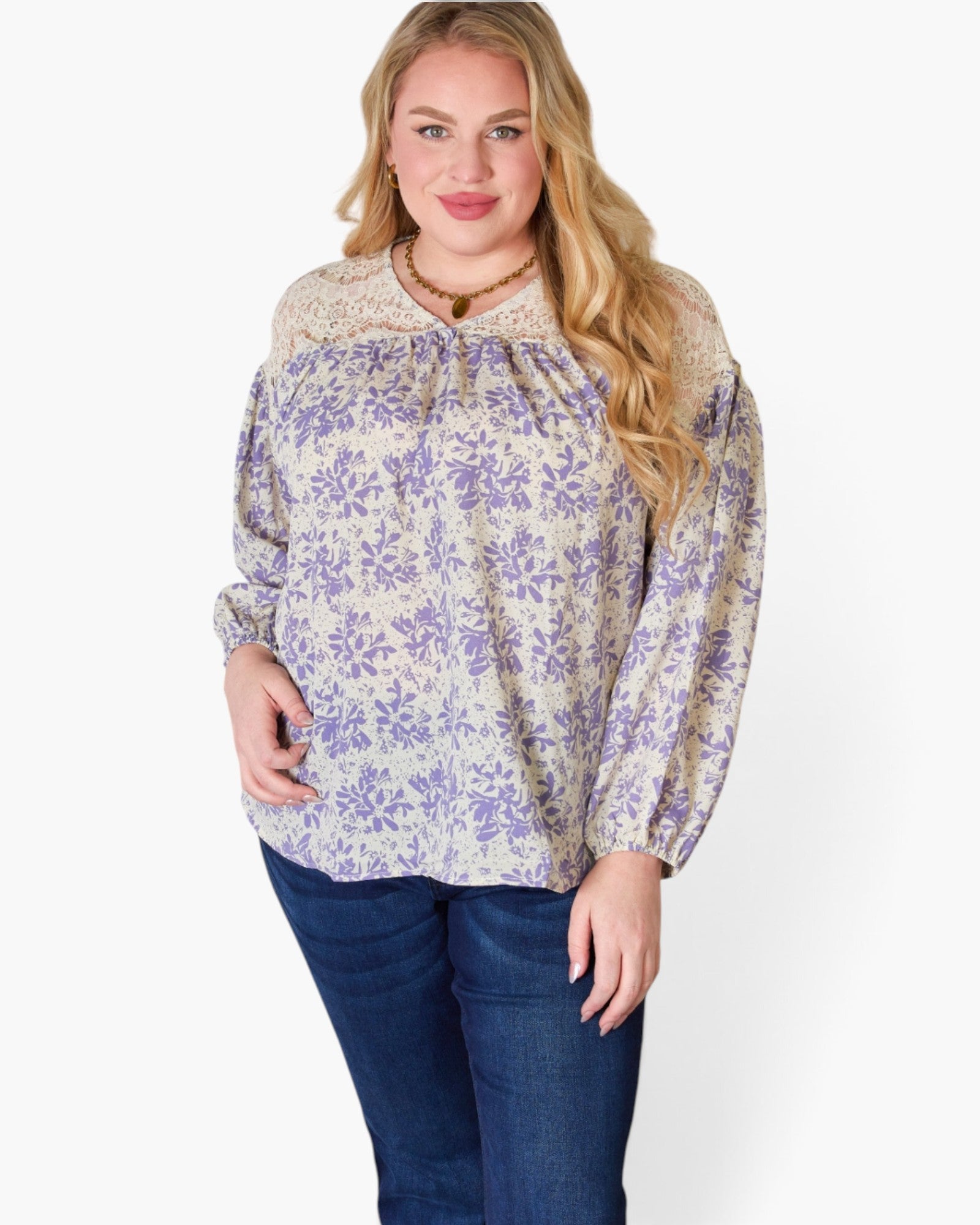 A woman modeling a long-sleeve blouse with a purple floral pattern on a cream background, featuring a lace-trimmed round neckline and gathered sleeves, paired with dark blue jeans against a plain white studio background.