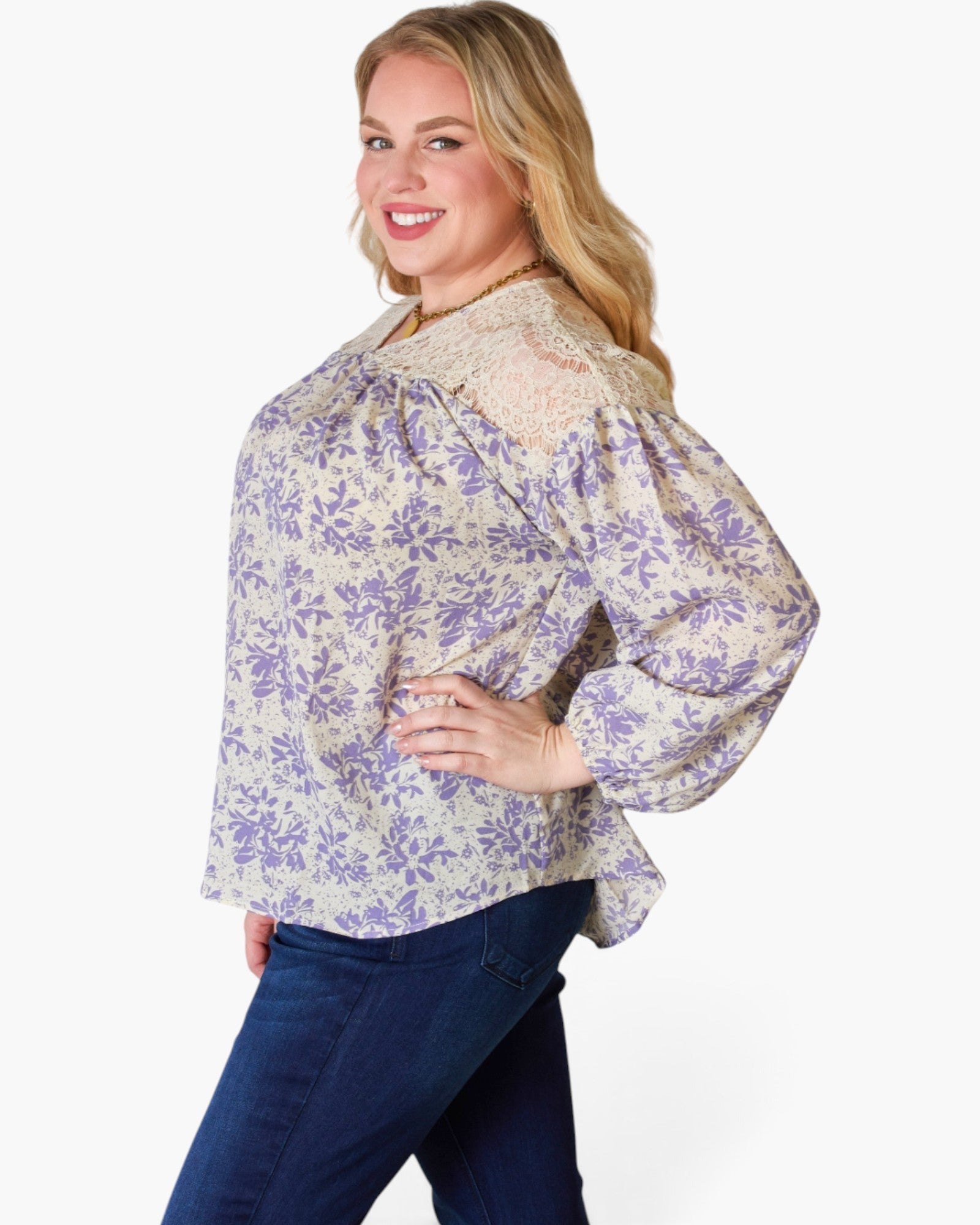 A woman modeling a long-sleeve blouse with a purple floral pattern on a cream background, paired with dark blue jeans, against a plain white studio background.