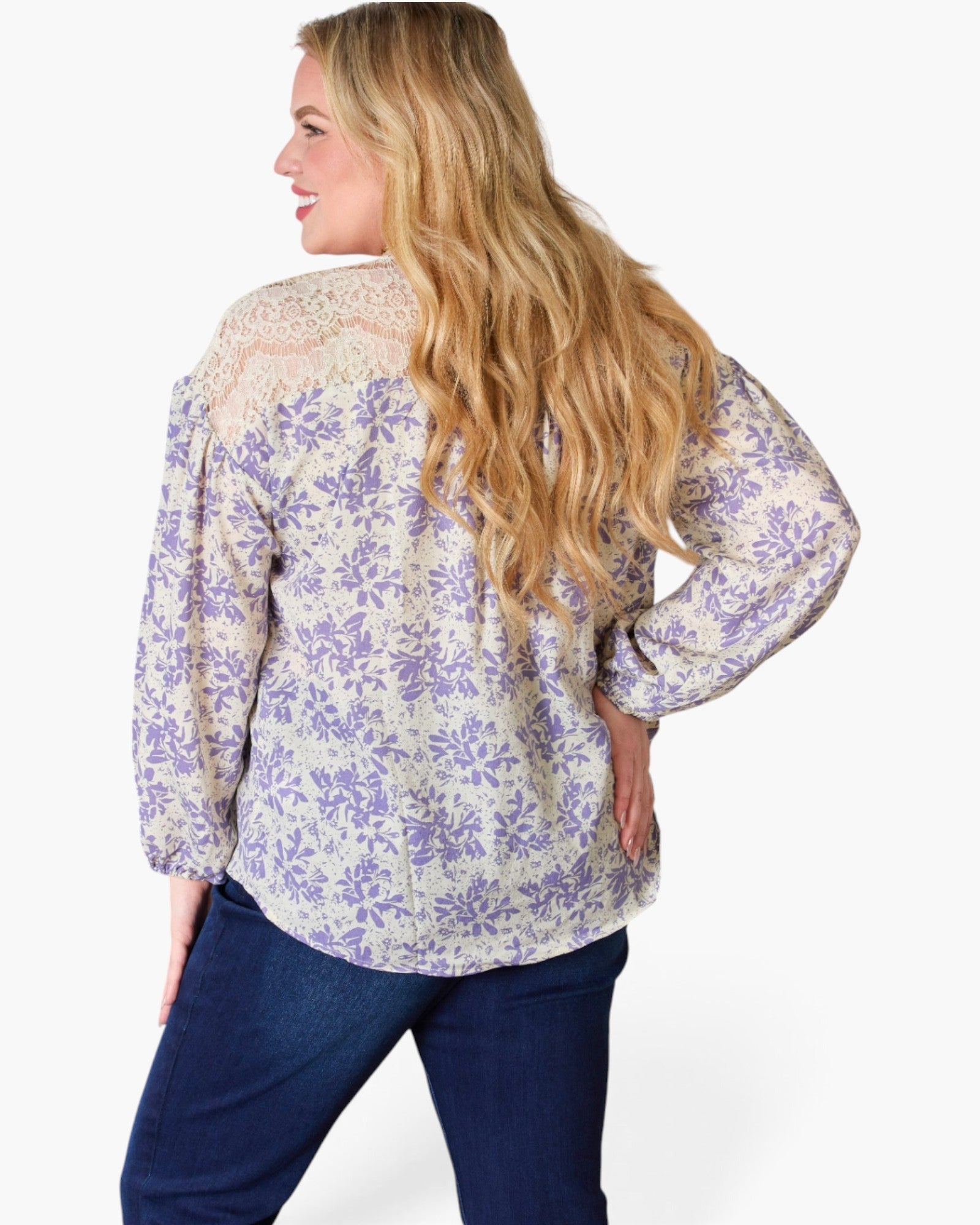 A woman shown from the back wearing a long-sleeve blouse with a purple floral pattern on a cream background, paired with dark blue jeans, against a plain white studio background.