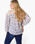 A woman shown from the back wearing a long-sleeve blouse with a purple floral pattern on a cream background, paired with dark blue jeans, against a plain white studio background.