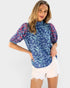 A woman modeling a short-sleeve mock-neck blouse with a blue, white, and pink floral pattern, paired with white denim shorts, against a light gray studio background.