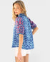 A woman modeling a short-sleeve blue floral shirt with puff sleeves, paired with white shorts, against a light gray studio background.