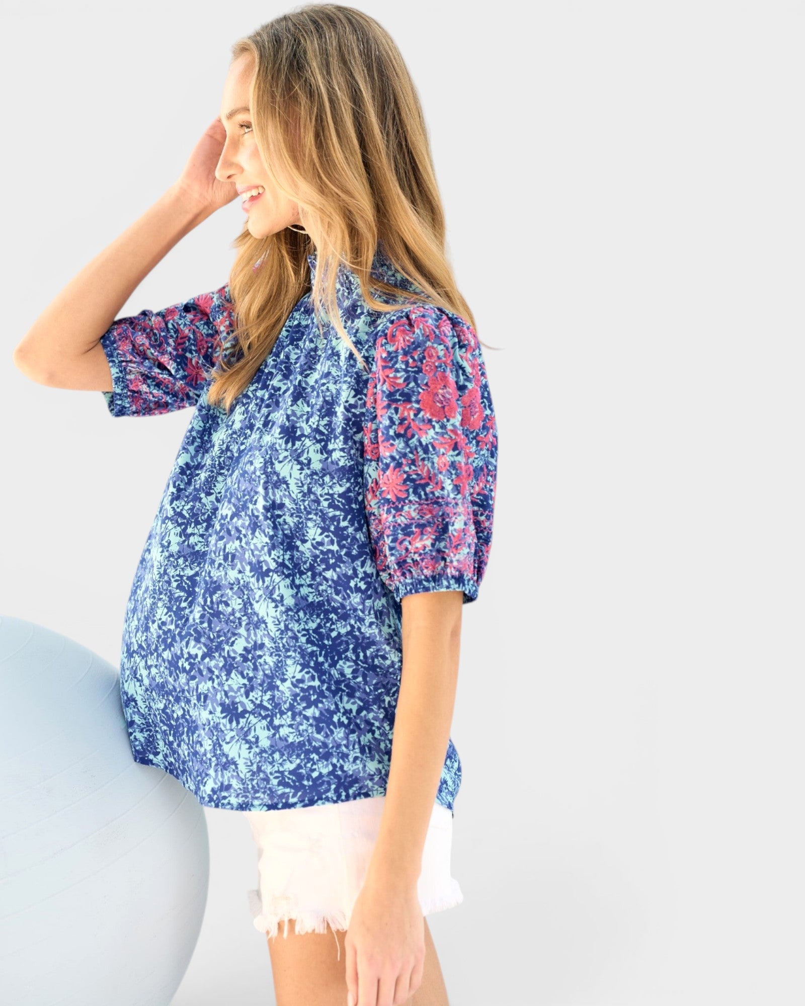 A pregnant woman wearing a short-sleeve maternity top with a blue, white, and pink floral pattern, paired with white shorts, posing against a light gray background.