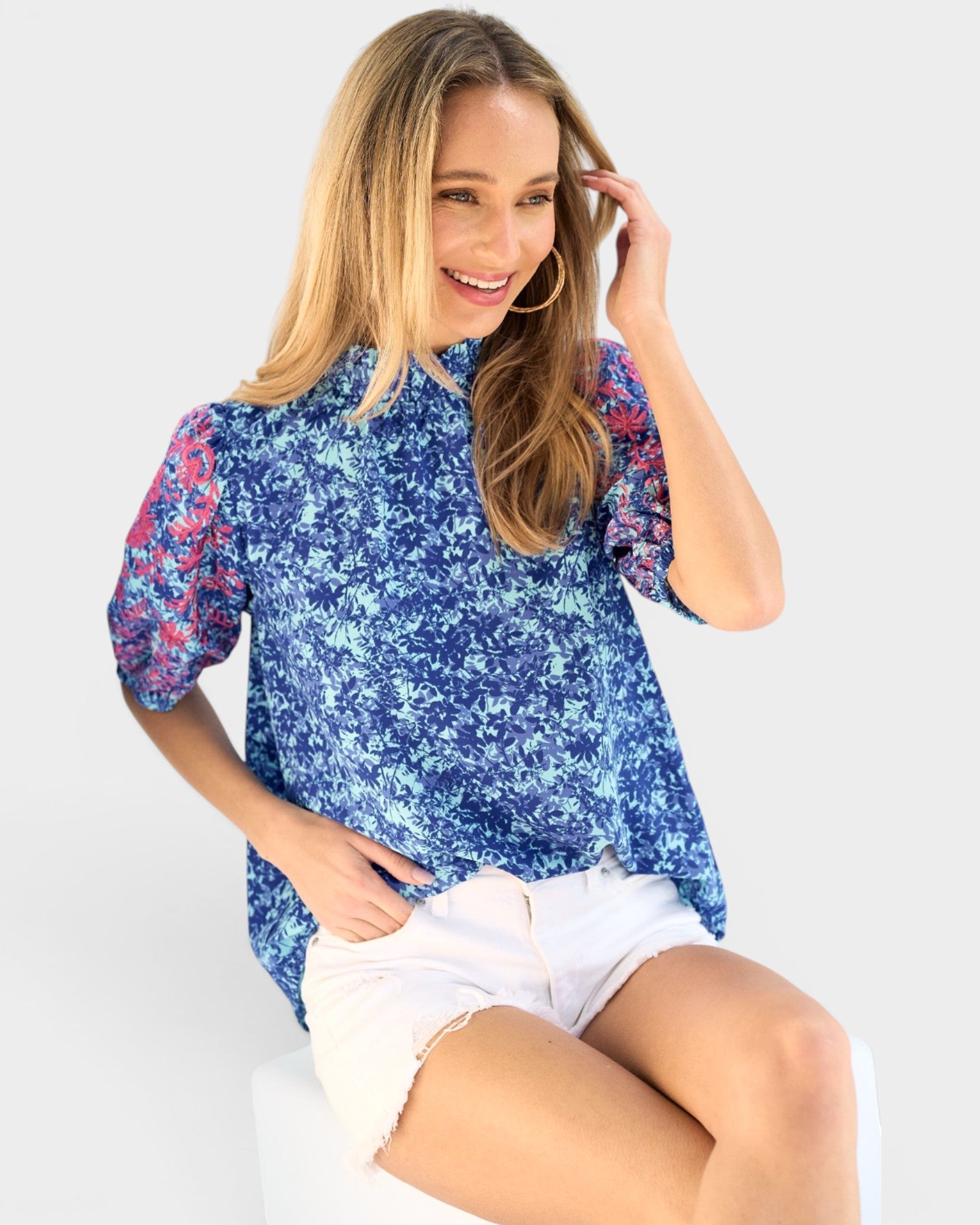 A woman wearing a blue floral short-sleeve blouse with pink and red patterned sleeves, paired with white shorts, seated against a light gray background.