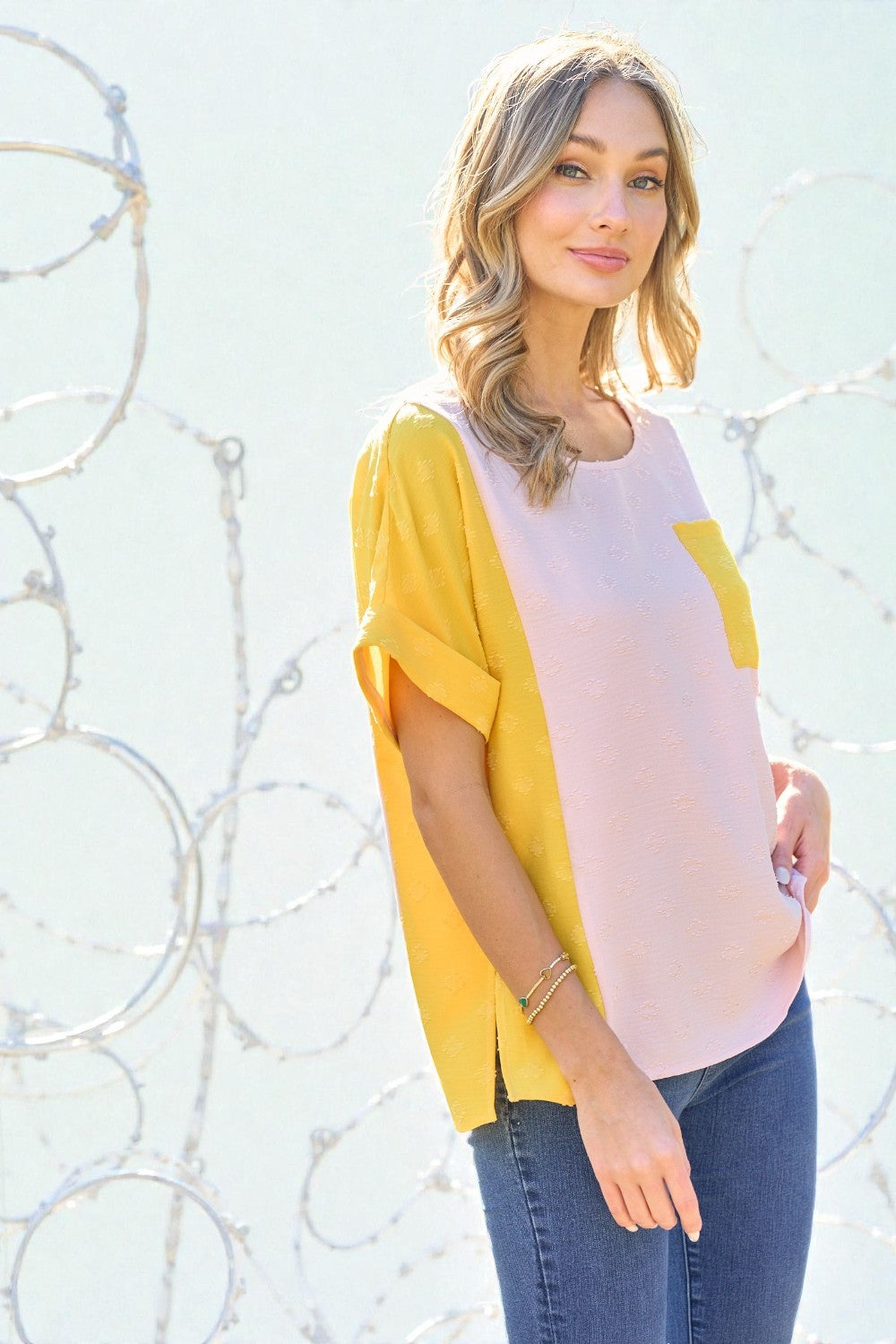 Woman wearing a yellow and pink top with blue jeans against a white wall.