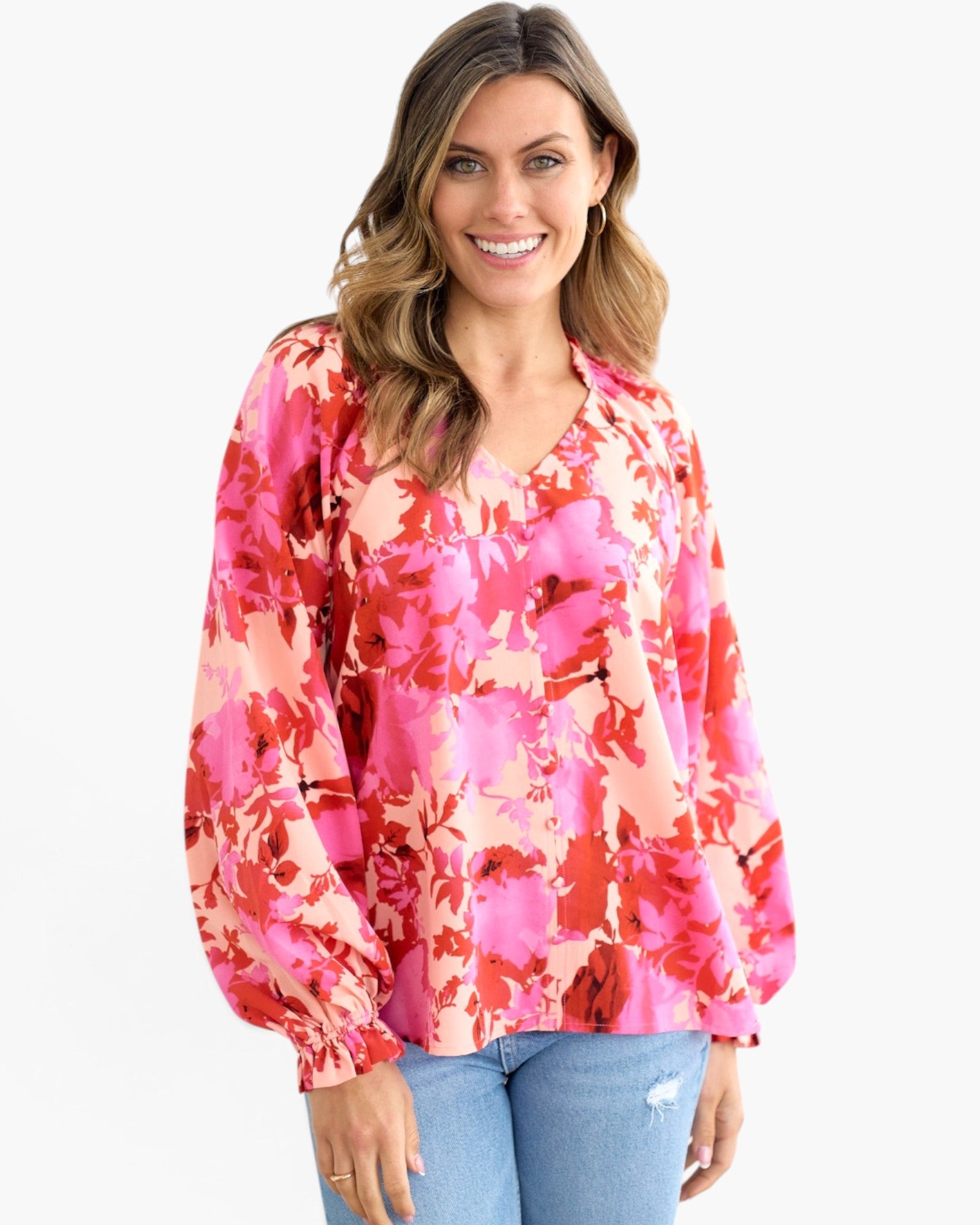 A woman wearing a vibrant pink and red floral long-sleeve blouse with a V-neckline, paired with light blue jeans, against a plain white background.