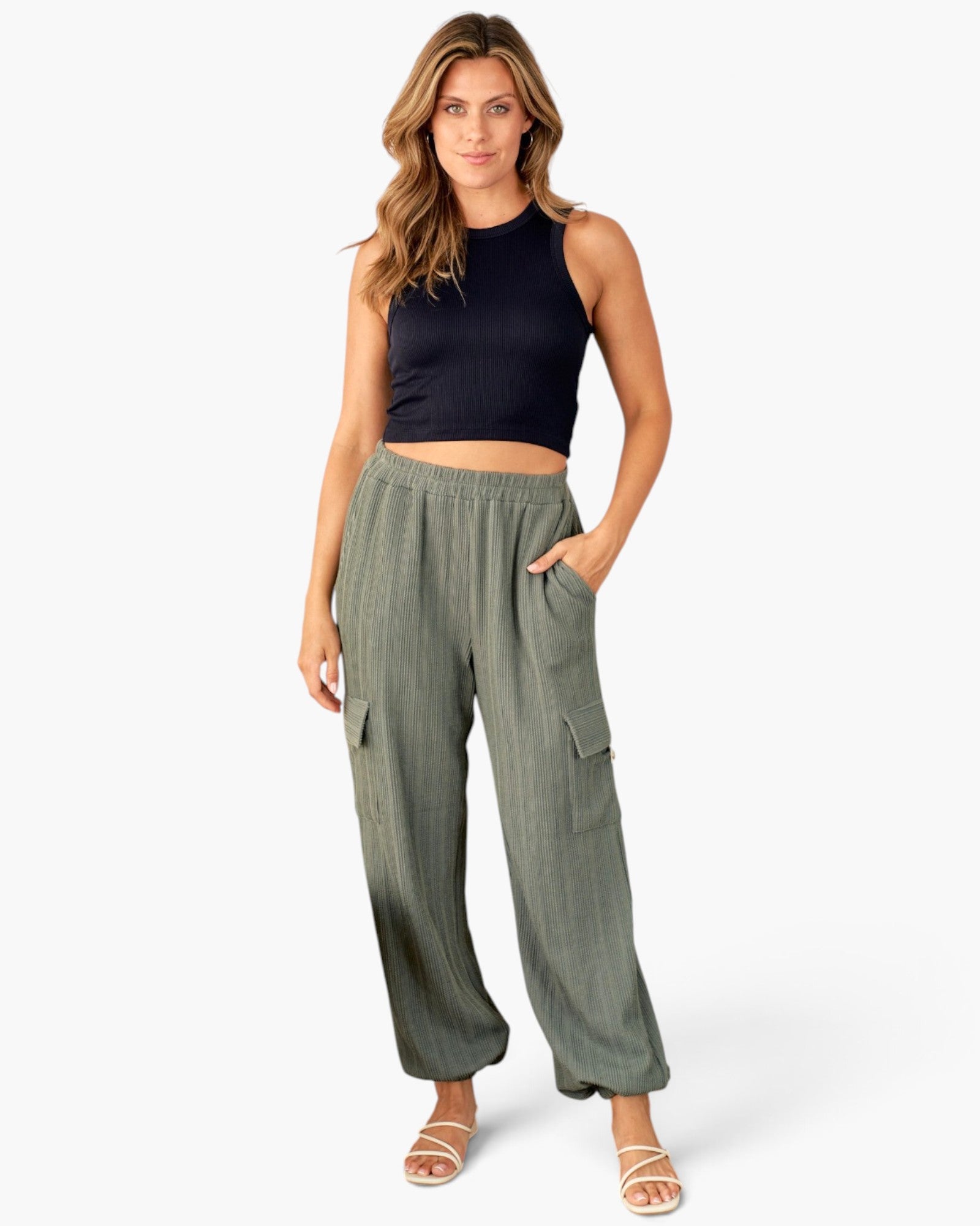 A woman modeling a sleeveless black crop top and olive green wide-leg cargo pants with side pockets, paired with beige strappy sandals, against a plain white studio background.