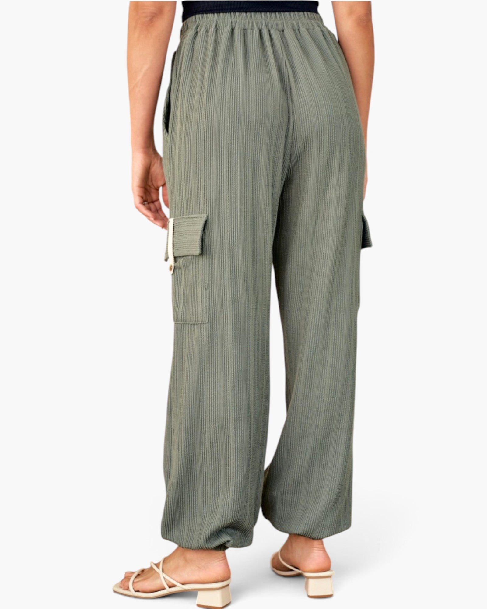 A person shown from the back wearing loose, wide-leg cargo pants in a muted sage green color with a subtle vertical textured stripe pattern. The pants feature an elastic waistband, side cargo pockets, and a relaxed, comfortable fit, paired with beige block-heel sandals against a clean white background.