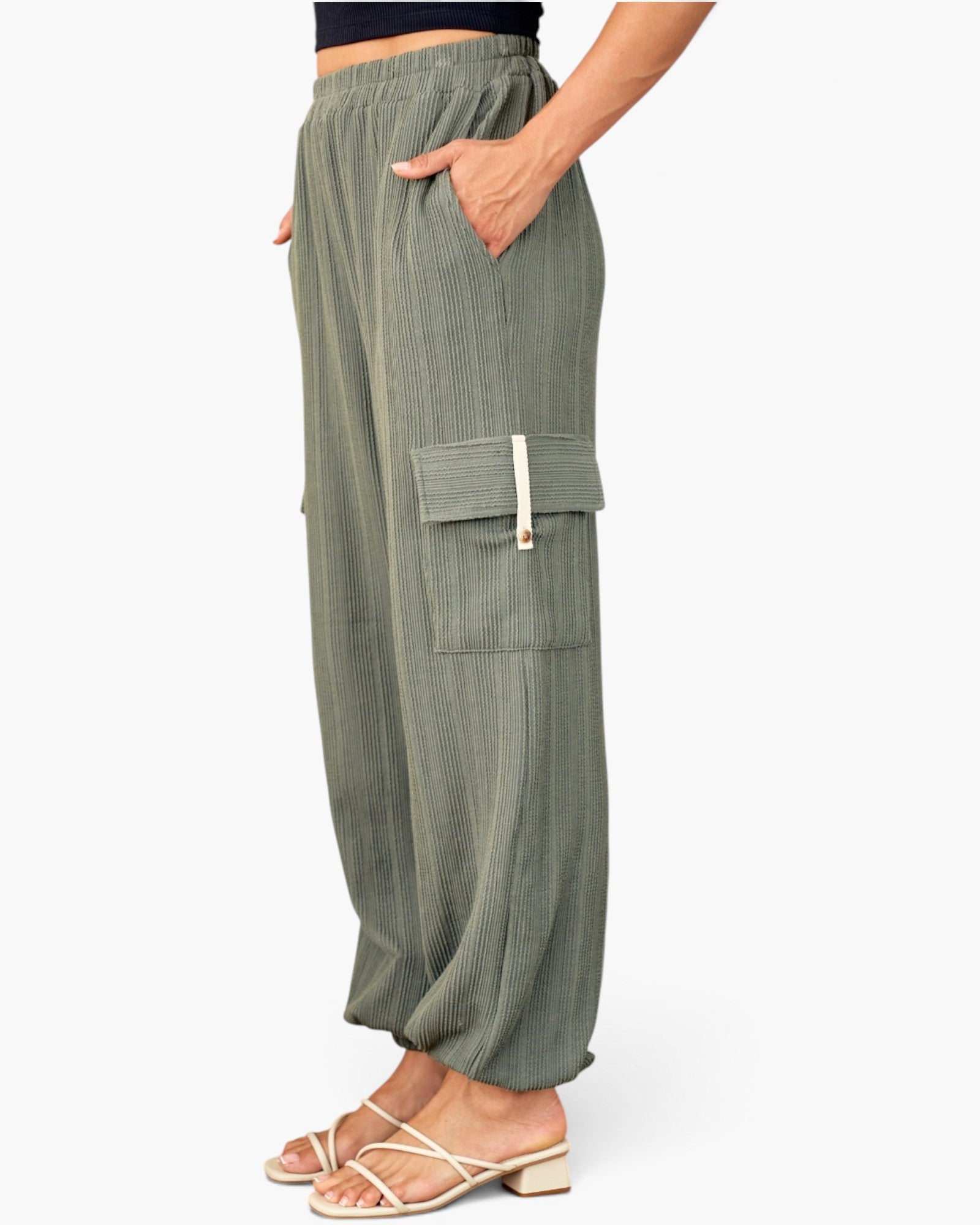 A person wearing loose olive green cargo pants with side flap pockets and white strappy heeled sandals, shown from the waist down against a light gray studio background.