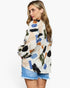 A woman wearing a long-sleeve blouse with an abstract multicolor brushstroke pattern on a cream base, paired with denim shorts, photographed against a plain white studio background.
