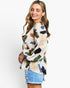 A woman modeling a long-sleeve blouse with an abstract multicolor brushstroke print on a cream base, paired with light blue denim shorts, against a plain white background.