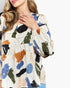 A woman wearing a long-sleeve blouse with an abstract brushstroke print in blue, green, orange, black, and beige on an off-white background.
