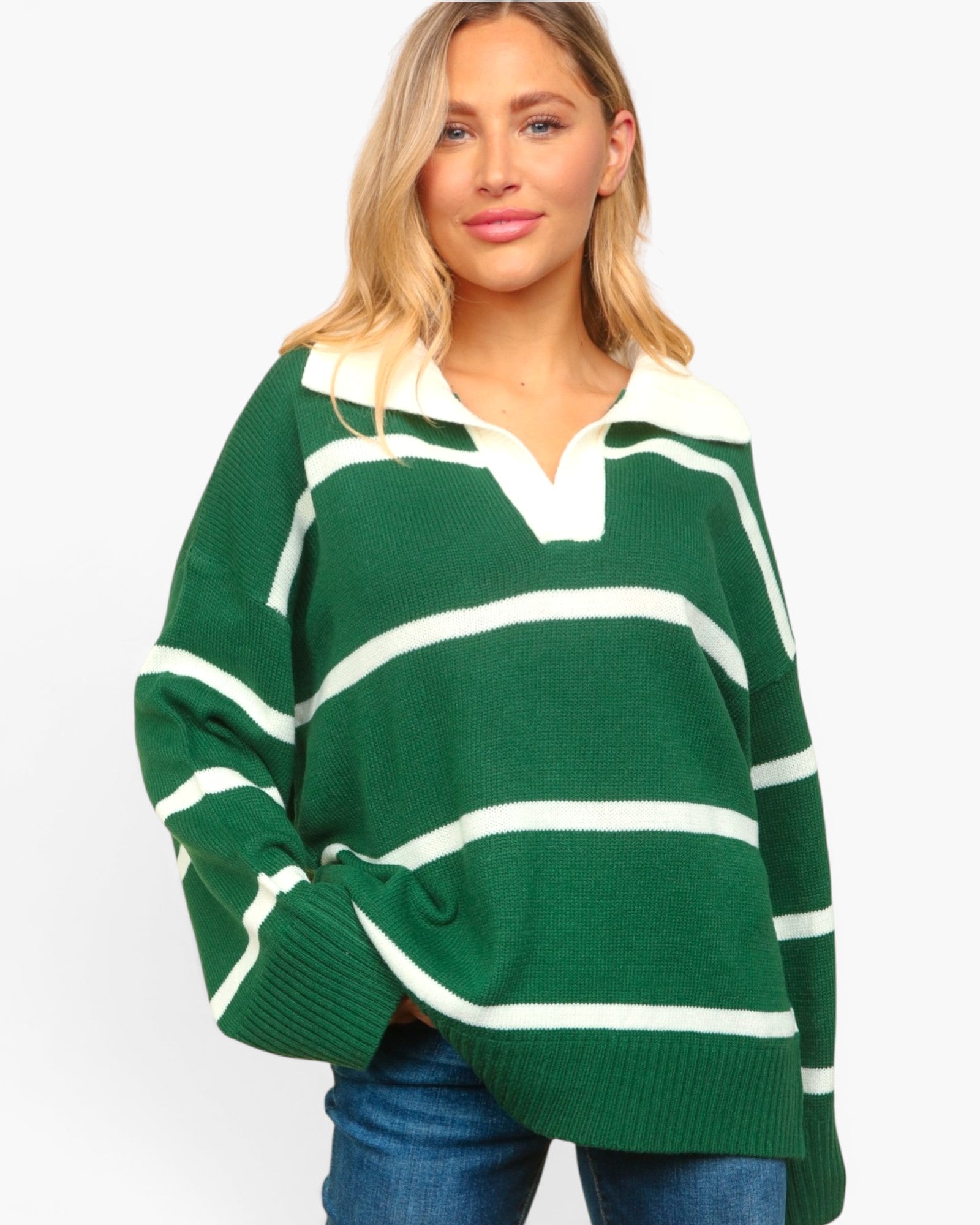 A woman wearing an oversized green and white striped knit rugby-style sweater with a split V-neck, paired with blue jeans, against a plain light gray background.