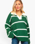 A woman wearing an oversized green and white striped knit rugby-style sweater with a split V-neck, paired with blue jeans, against a plain light gray background.