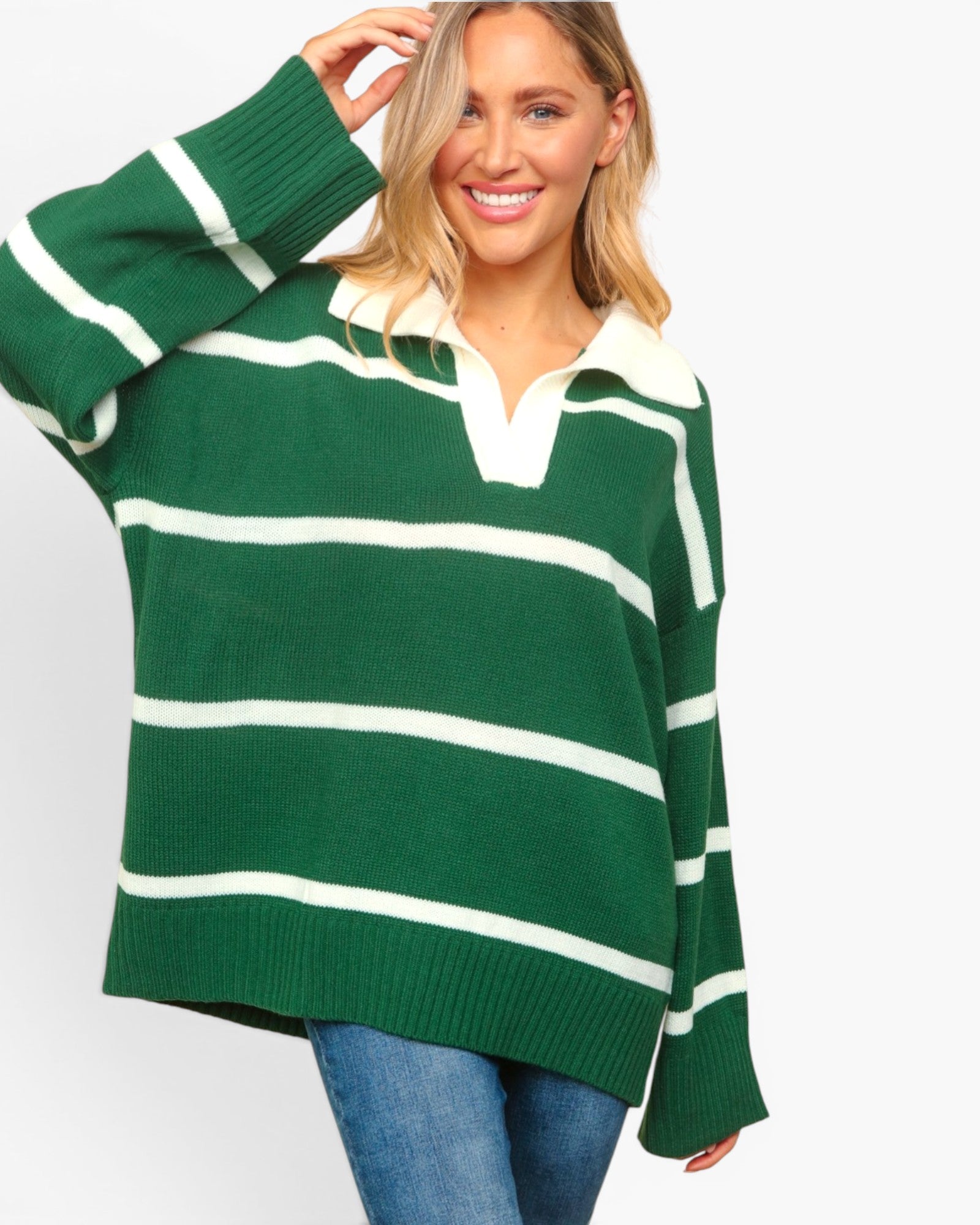 A woman wearing an oversized green and white striped knit sweater with a polo-style collar, paired with blue jeans, against a plain light gray background.