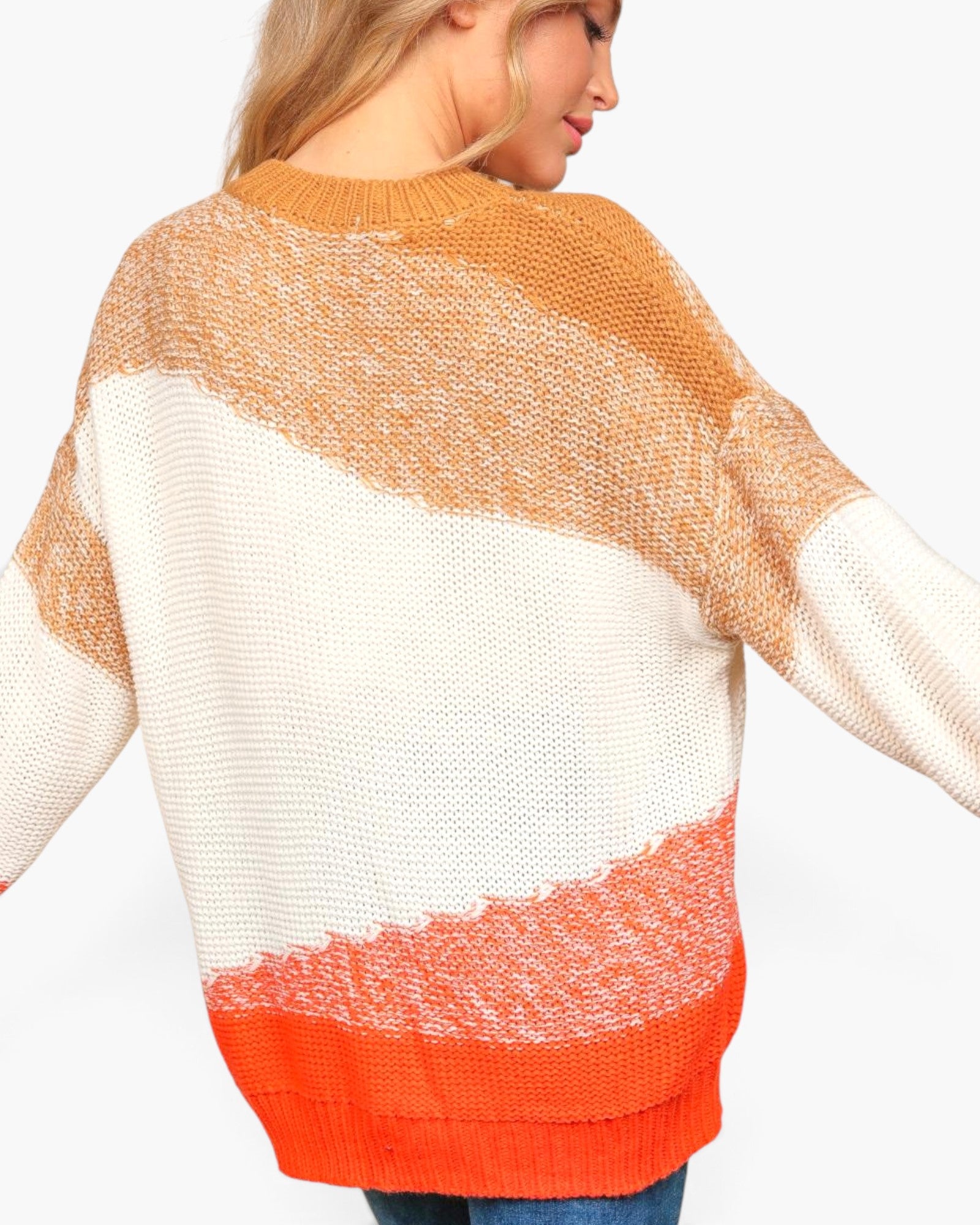 A woman wearing a chunky knit sweater with diagonal color-blocked stripes in tan, off-white, and bright orange, shown from a side-back angle against a plain white background.