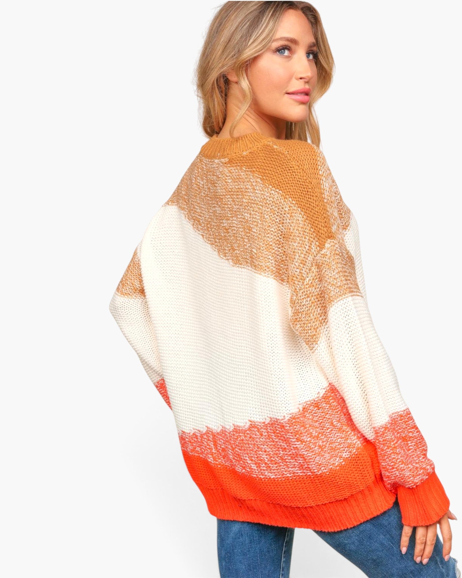 A woman wearing a chunky knit sweater with large color-blocked stripes in cream, tan, and bright orange, paired with blue jeans, shown from a three-quarters back view against a plain white studio background.