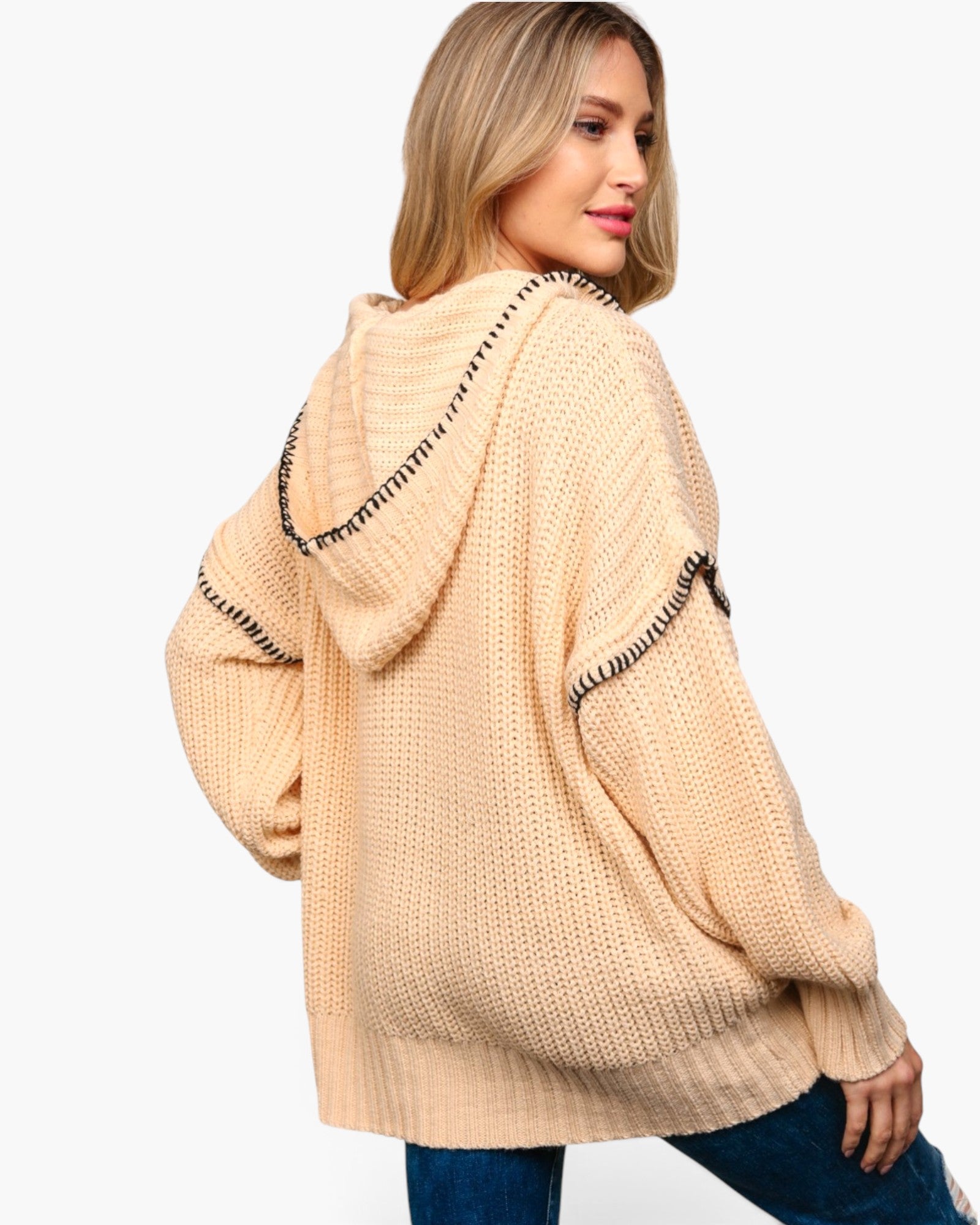 A woman wearing a chunky knit hooded sweater in a light peach-beige color with black embroidered trim details, shown from a three-quarter back view against a plain white background.