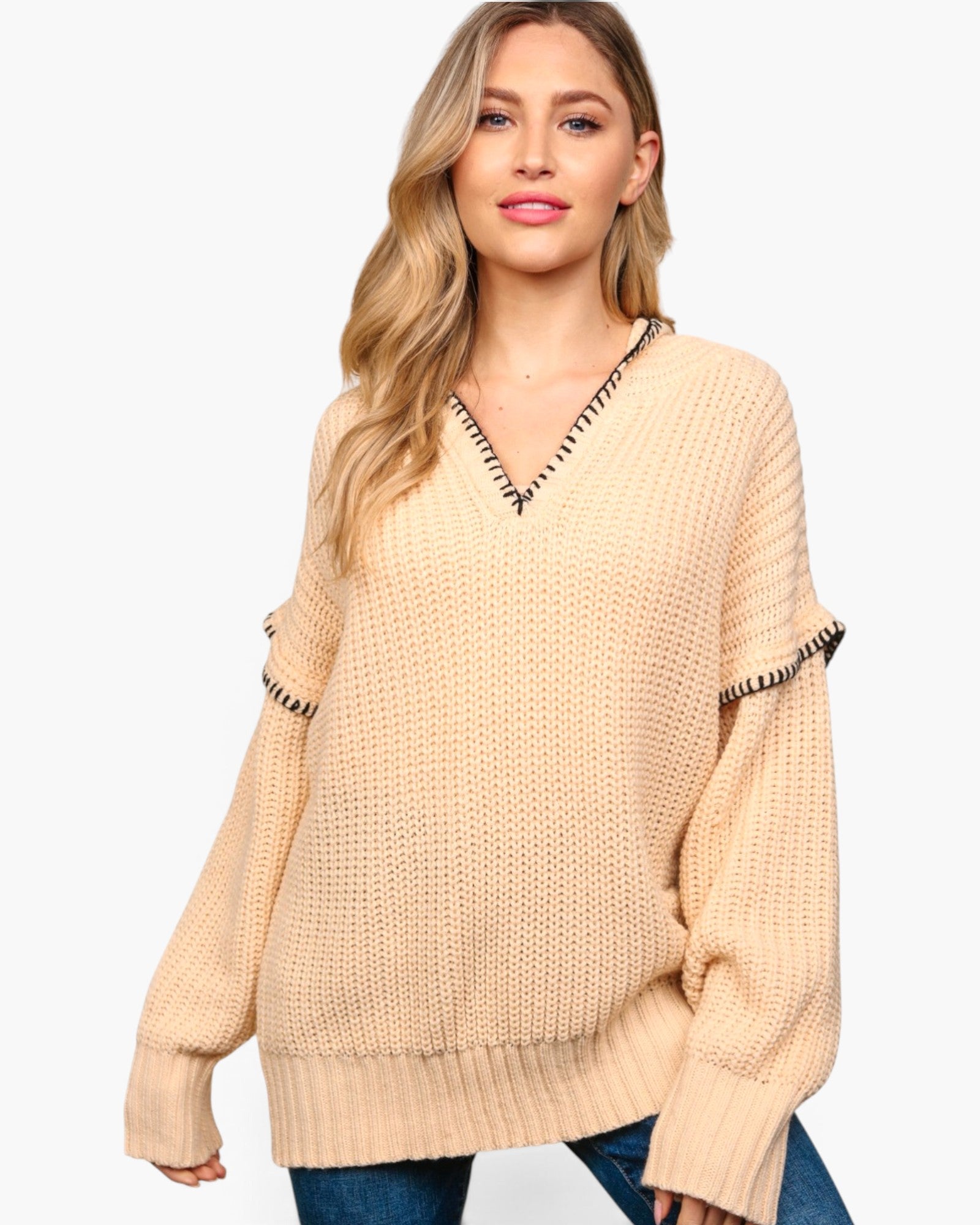 A woman wearing a beige chunky knit sweater with black and white striped trim along the V-neck and sleeve cuffs, paired with blue jeans, against a plain white background.