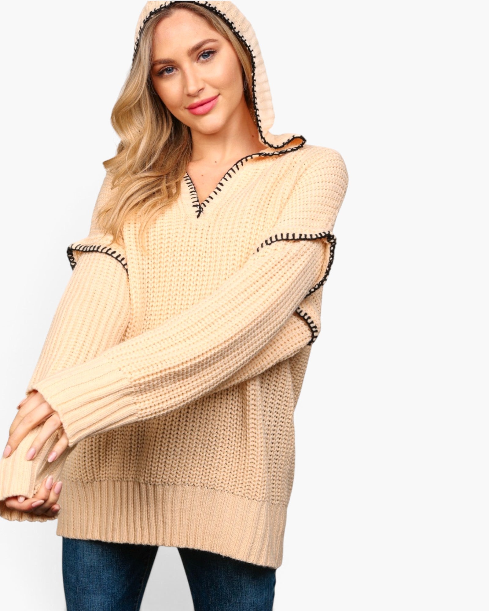 A woman wearing a chunky knit hooded sweater in a light beige color with black and white striped trim details along the hood, neckline, and sleeve cuffs, paired with blue jeans against a plain white background.