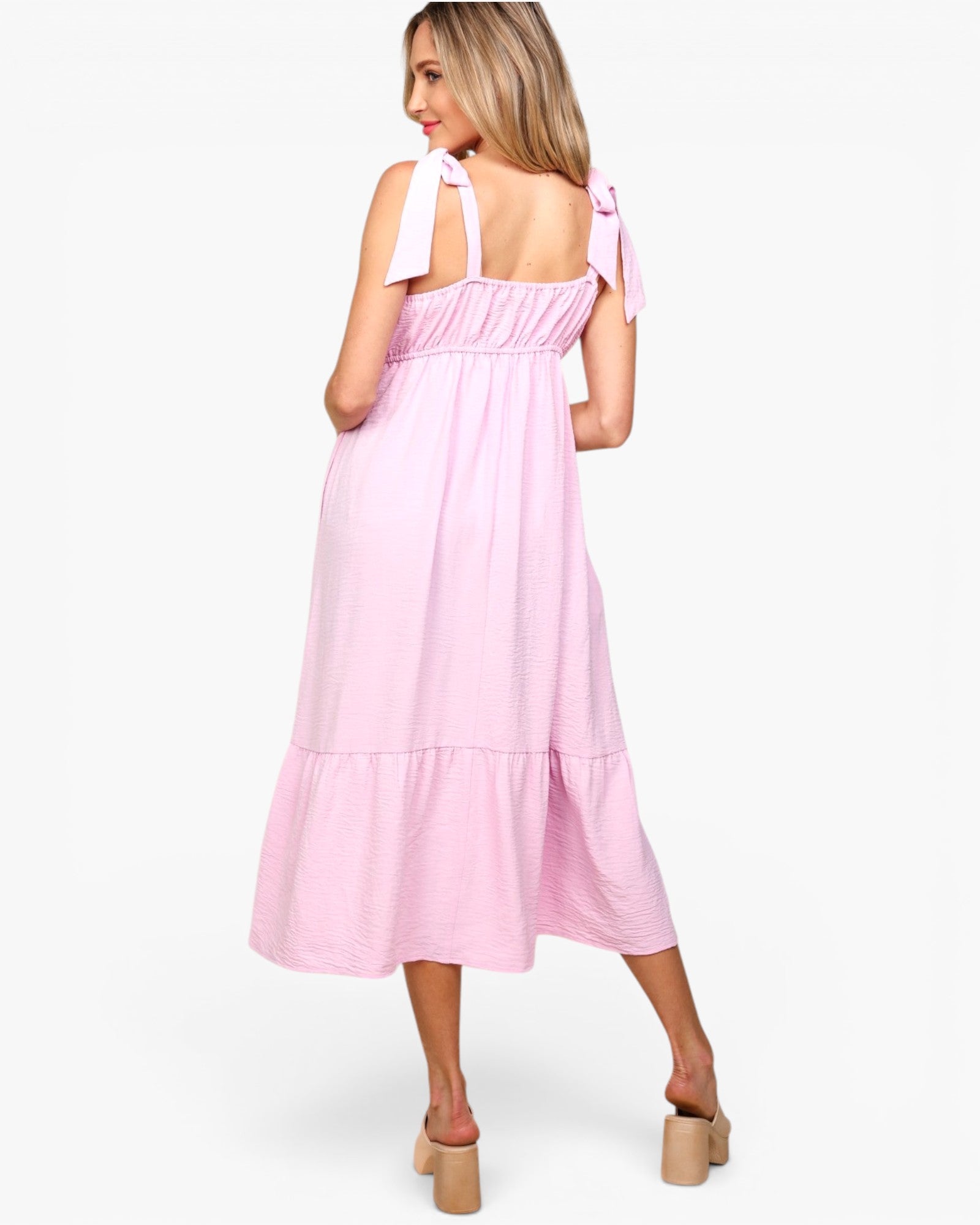 A woman modeling a light pink tiered midi dress with tie-shoulder straps, shown from the back against a plain white studio background.