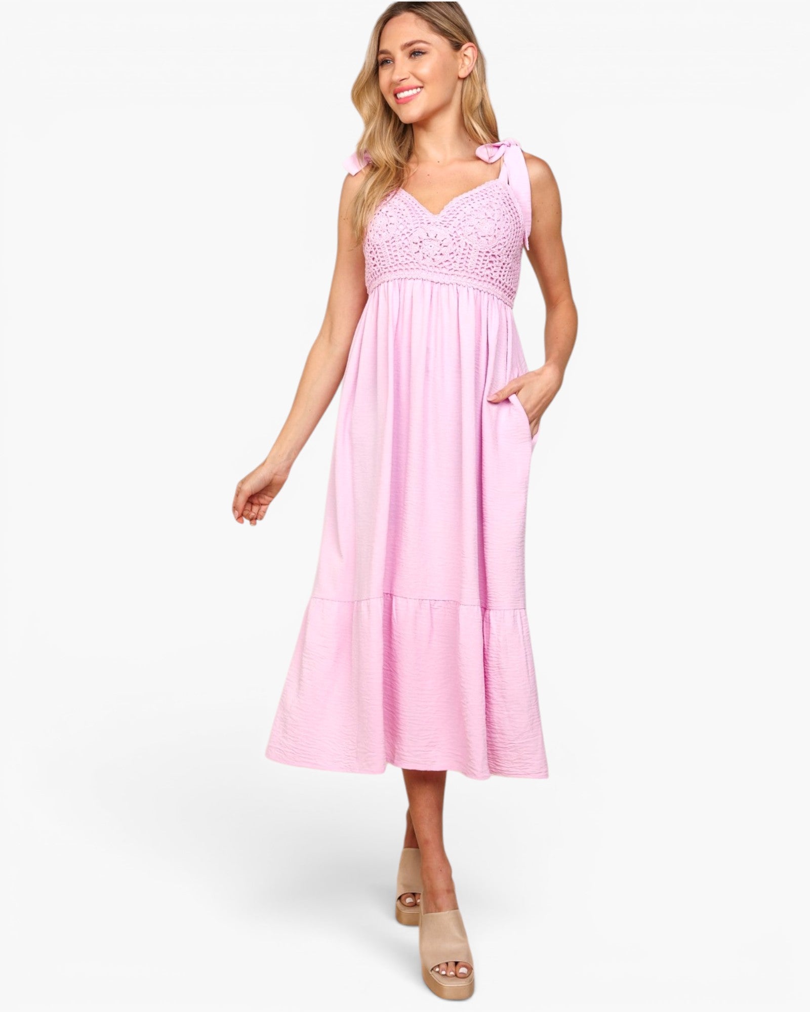 A woman modeling a light pink sleeveless midi dress with a lace bodice, shoulder tie details, and side pockets, paired with beige platform sandals against a plain white background.