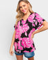 A woman wearing a black short-sleeve V-neck top with a vibrant pink floral print, paired with denim shorts, against a light gray studio background.
