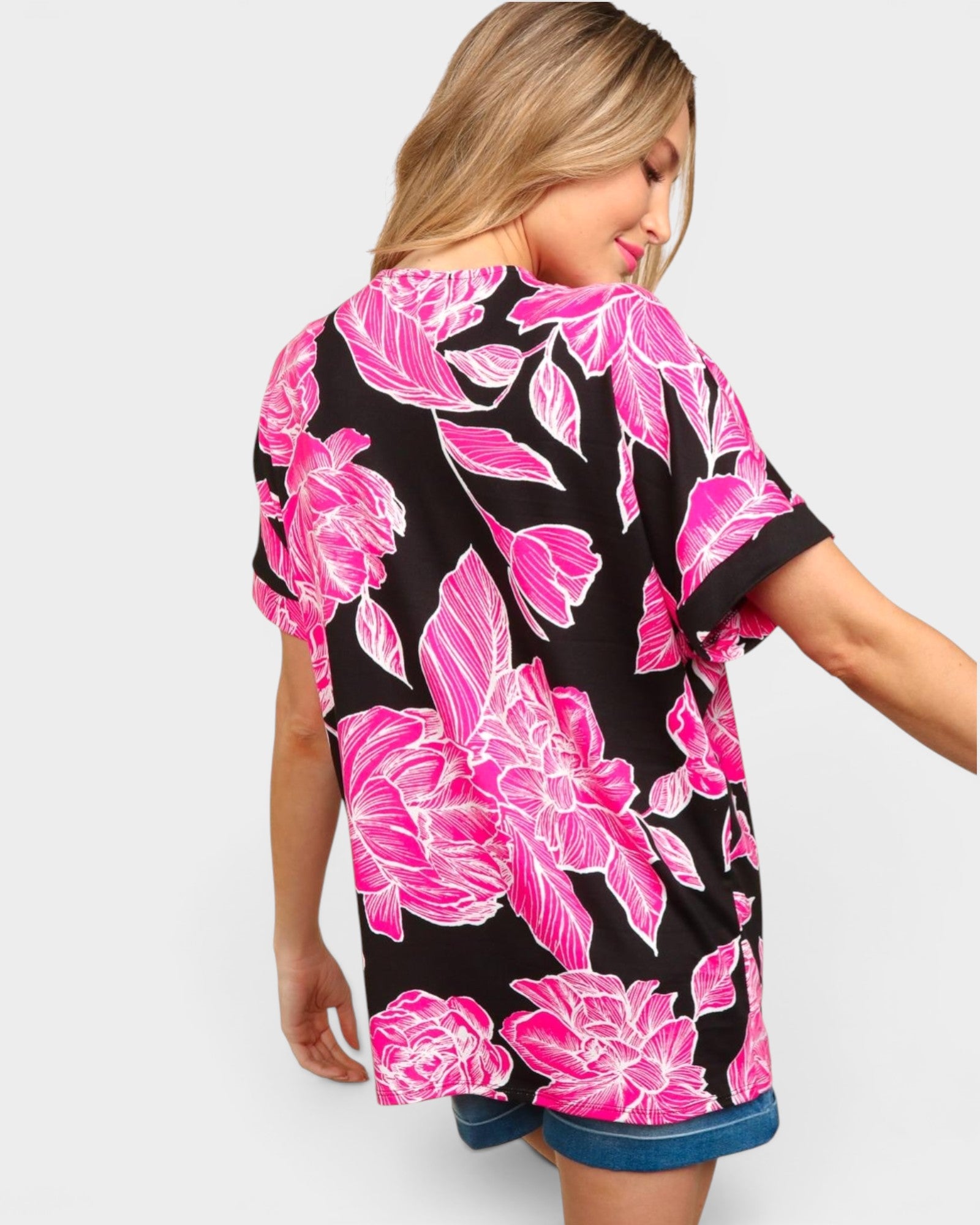 A woman wearing a black short-sleeve t-shirt with a vibrant pink floral print, shown from a three-quarters back view against a light gray background.