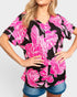 A woman wearing a black short-sleeve V-neck top with a bold hot pink floral print, paired with denim shorts, against a light gray background.