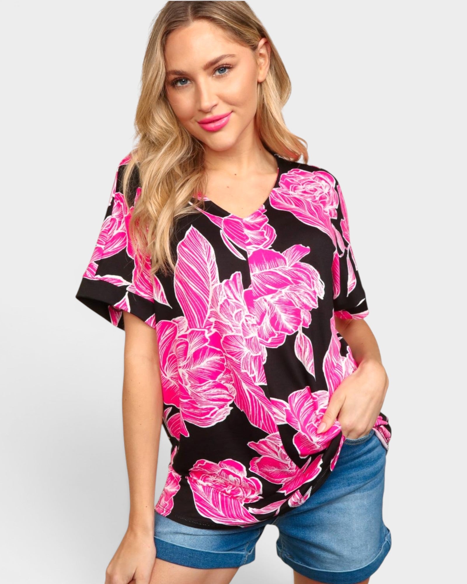 A woman modeling a black short-sleeve V-neck top with a vibrant pink and white floral print, paired with denim shorts, against a light gray studio background.