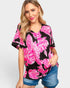 A woman modeling a black short-sleeve V-neck top with a vibrant pink and white floral print, paired with denim shorts, against a light gray studio background.