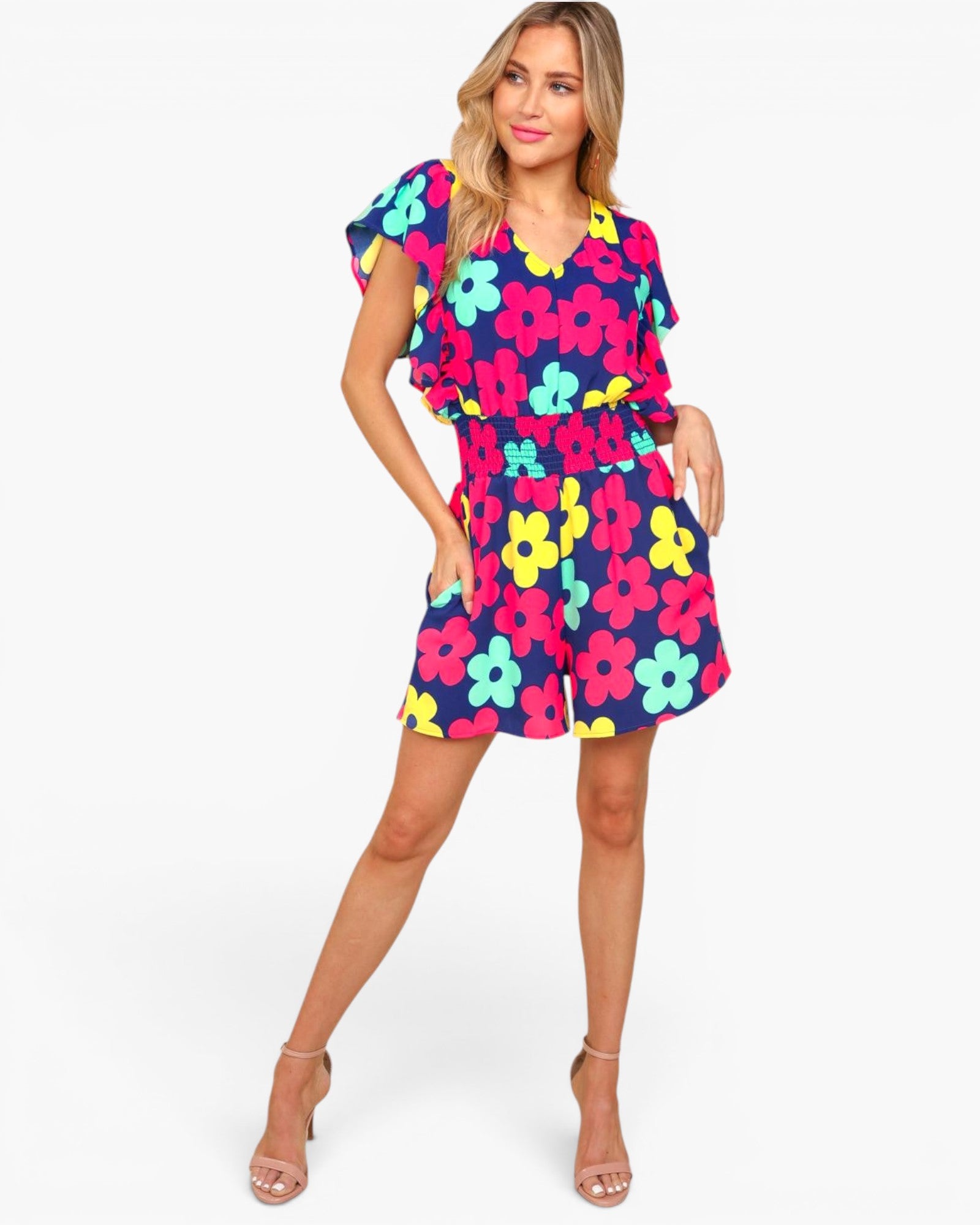 A woman modeling a short romper with a colorful floral print on a solid white background. The romper features a navy blue base with bright pink, yellow, and light blue flowers, and is styled with clear high-heeled sandals.