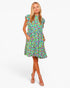 A woman modeling a short-sleeve knee-length dress with a colorful floral print on a light green background, paired with beige high-heeled sandals, against a plain white studio background.