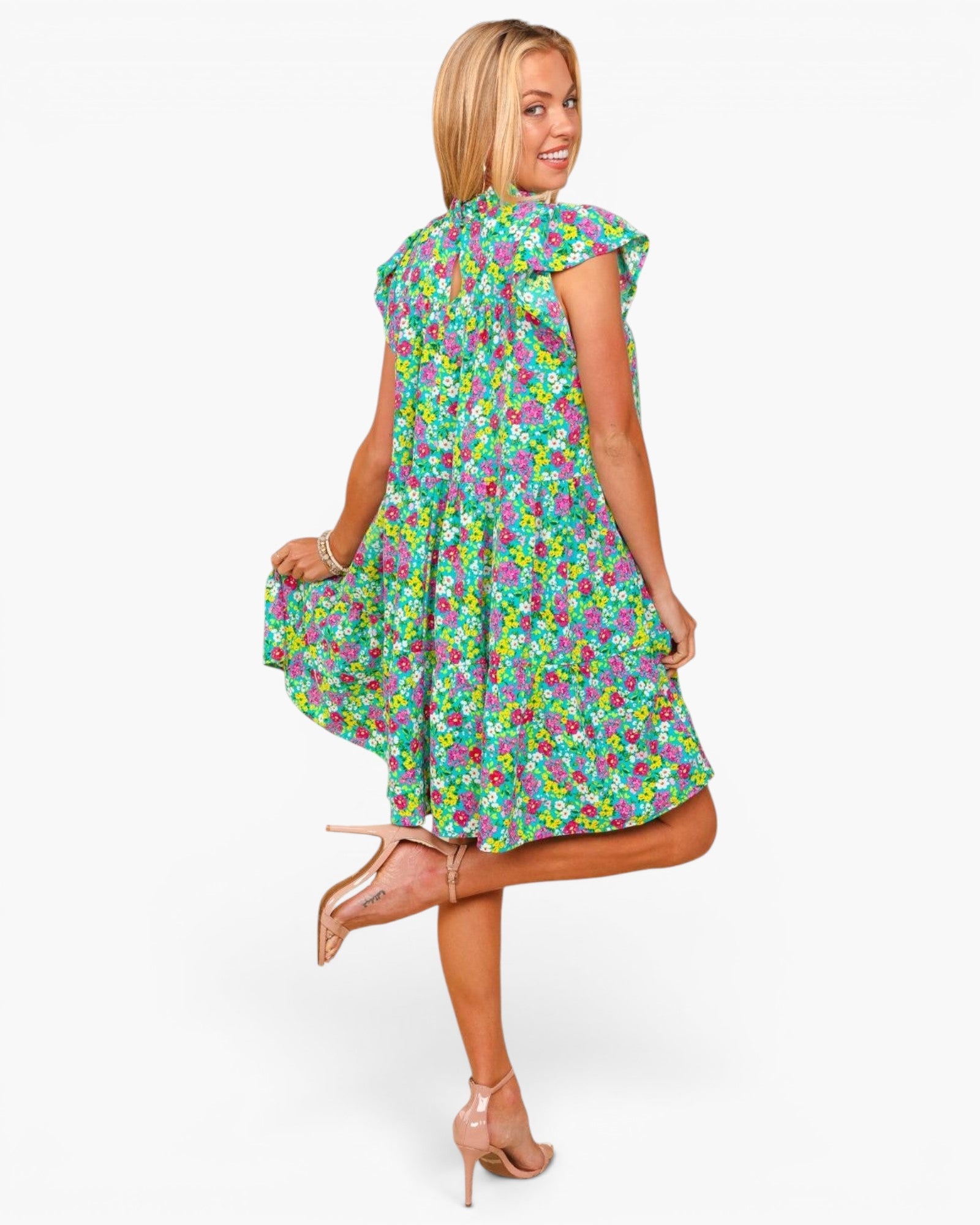 A woman modeling a sleeveless high-low dress with a colorful floral print, shown from a back-three-quarter angle against a plain white studio background.