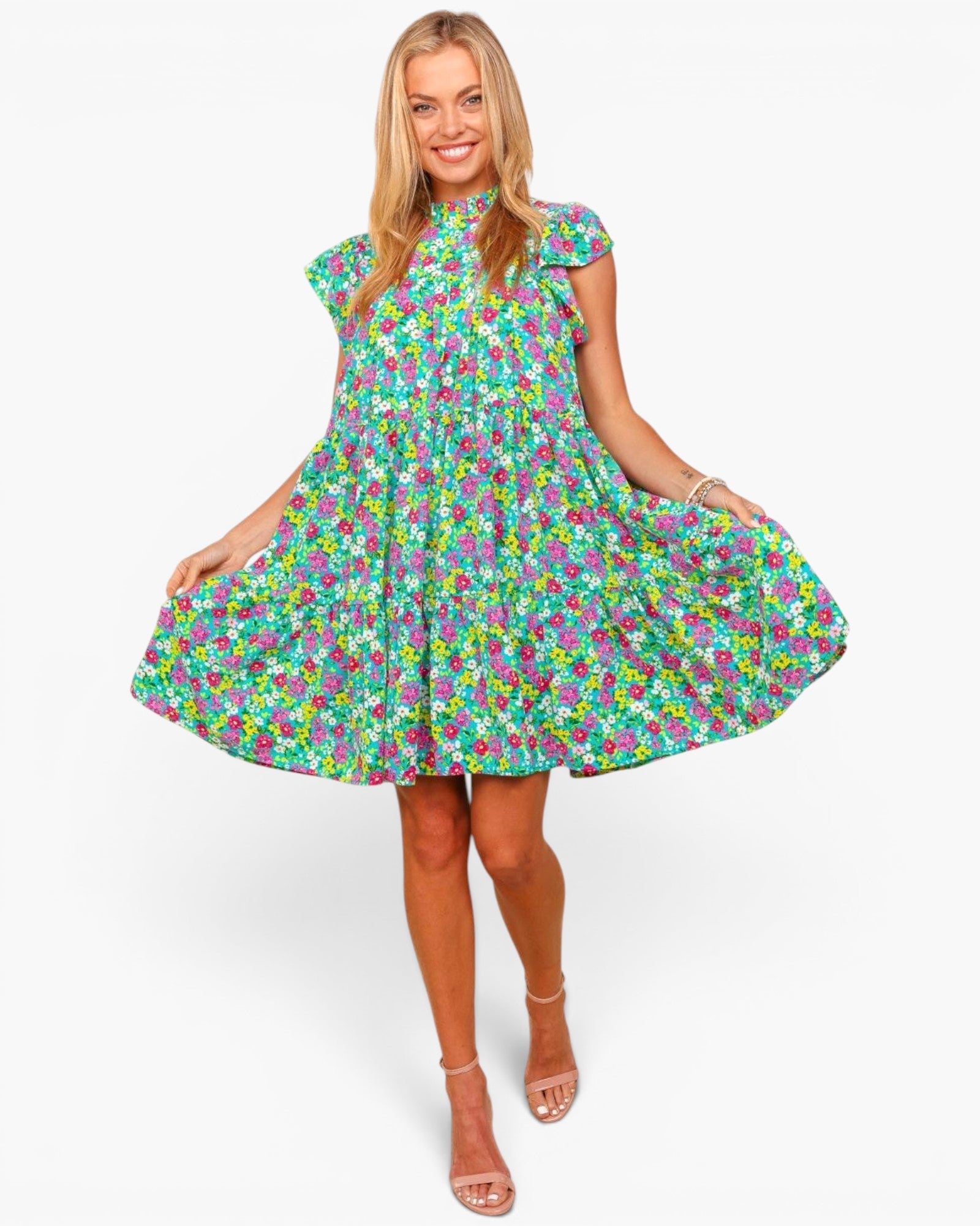 A woman modeling a short-sleeve, high-neck, flared mini dress with an all-over colorful floral print on a light green background, paired with beige strappy high-heeled sandals against a plain white studio background.