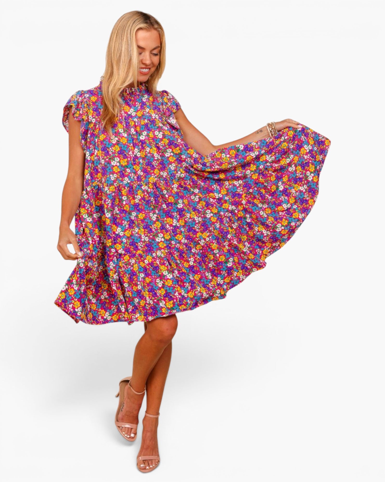 A woman modeling a short-sleeve, flowy A-line dress with an all-over colorful floral print. She is holding out the side of the dress to show its volume, set against a plain white studio background.