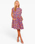A woman modeling a short, sleeveless A-line dress with a vibrant multicolor floral print in purple, pink, yellow, and blue. The dress features ruffled cap sleeves, a round neckline, and side pockets, shown against a plain white studio background.