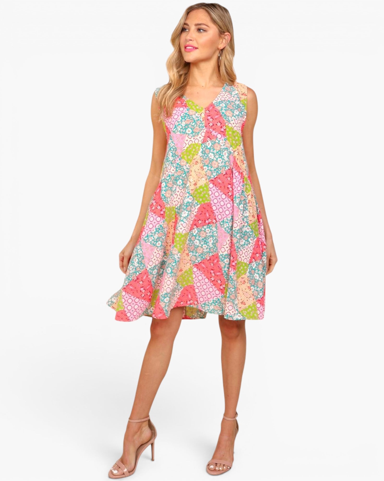 A woman modeling a sleeveless V-neck A-line dress with a colorful patchwork triangle print in pink, teal, green, and white, paired with nude high-heeled sandals against a light gray studio background.