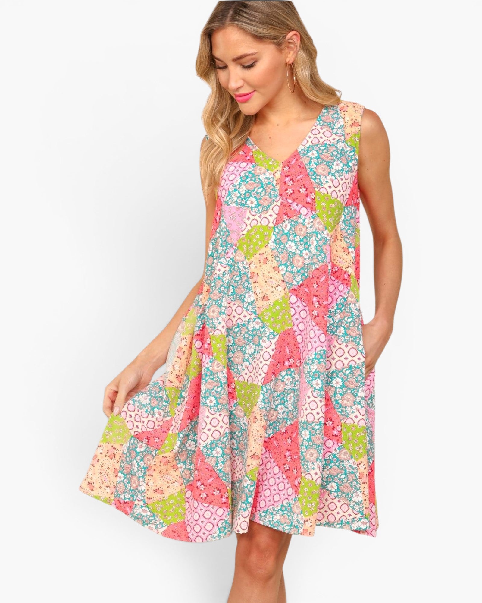 A woman modeling a sleeveless V-neck A-line dress with a colorful patchwork floral print in pink, teal, green, and cream on a light gray background.