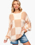 A woman wearing a chunky knit sweater with a large checkered pattern in beige and cream, paired with blue denim shorts, against a plain white background.