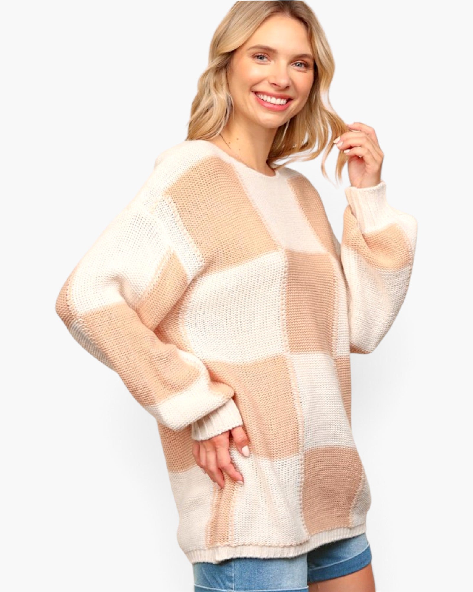 A woman wearing a chunky knit patchwork sweater with large beige and cream blocks, paired with blue denim shorts, against a plain white background.